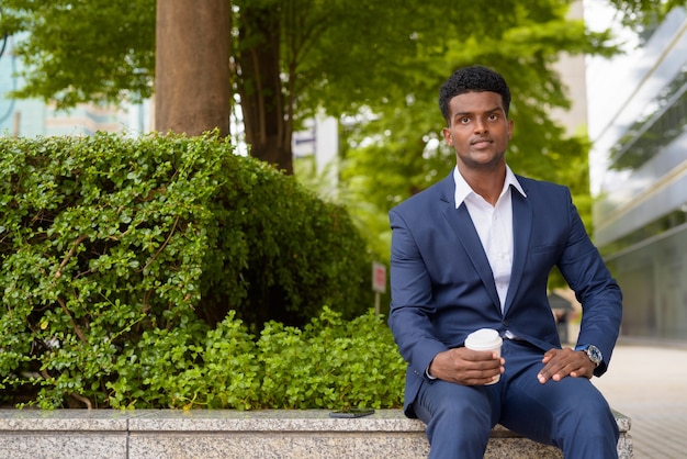 Portrait de jeune homme d'affaires africain tenant une tasse de café à emporter à l'extérieur de la ville tout en étant assis, prise de vue horizontale