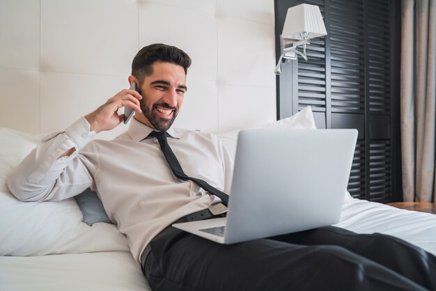 Portrait de jeune homme d'affaires allongé sur le lit et parler au téléphone tout en travaillant sur son ordinateur portable dans la chambre d'hôtel. Concept de voyage d'affaires.