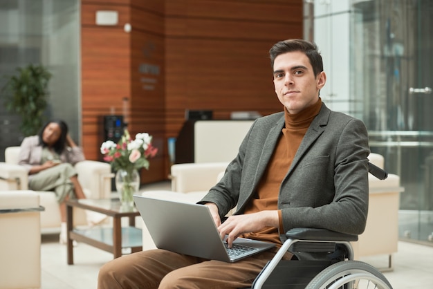 Portrait de jeune homme d'affaires assis dans un fauteuil roulant regardant la caméra et tapant sur un ordinateur portable, il travaille en ligne