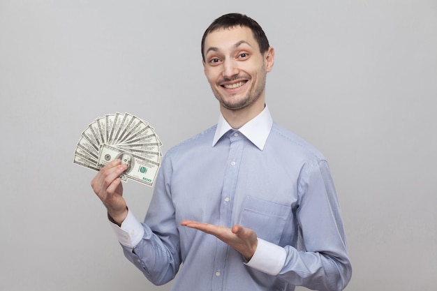 Portrait d'un jeune homme d'affaires attrayant et satisfait en chemise bleue debout, donnant et pointant avec un fan d'argent et un sourire à pleines dents. Prise de vue en studio intérieur, isolé, fond gris, espace pour copie