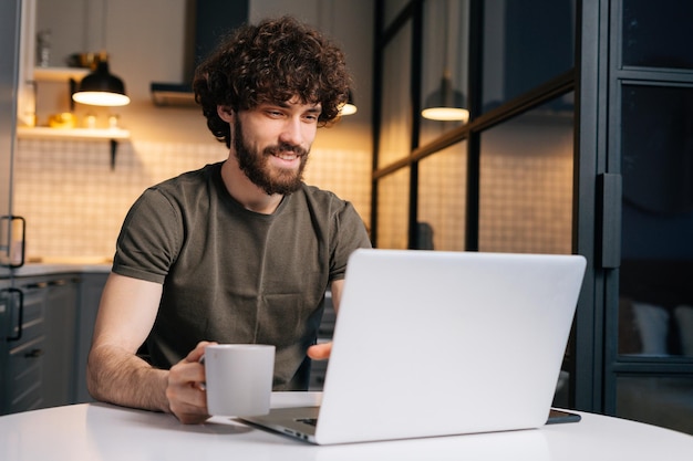 Portrait d'un jeune homme d'affaires barbu concentré tenant dans la main une tasse avec le café du matin assis à table avec un ordinateur portable regardant à l'écran