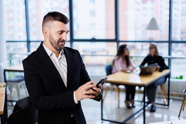 Portrait de jeune homme d'affaires élégant debout dans un grand bureau moderne au dernier étage et regardant le téléphone