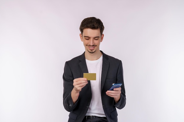 Portrait d'un jeune homme d'affaires heureux debout à l'aide d'un téléphone portable et détenant une carte bancaire de crédit isolée sur fond blanc studio de fond