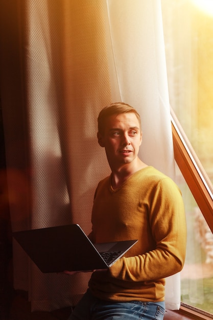 Portrait de jeune homme d'affaires avec ordinateur portable à la fenêtre du salon dans la maison de campagne. Homme bourreau de travail dans des vêtements décontractés à la maison travaillant en vacances. Inspiration créative et création d'entreprise. Espace de copie