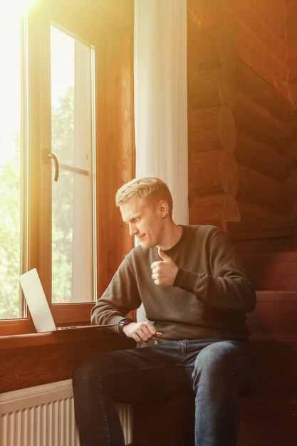 Portrait de jeune homme d'affaires avec ordinateur portable à la fenêtre du salon dans la maison de campagne. Homme bourreau de travail dans des vêtements décontractés à la maison travaillant en vacances. Inspiration créative et création d'entreprise. Espace de copie