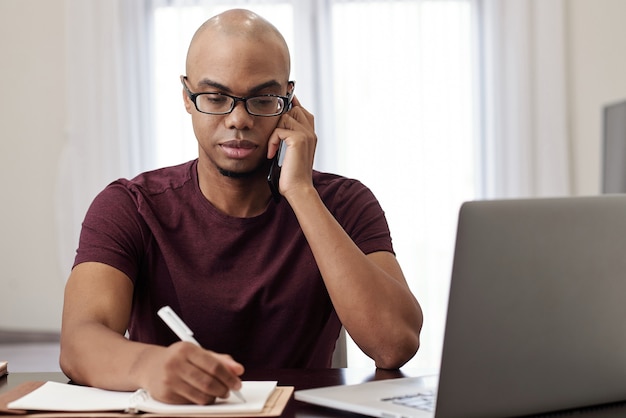 Portrait de jeune homme d'affaires sérieux appelant au téléphone et prenant des notes dans le planificateur en parlant au téléphone avec le client