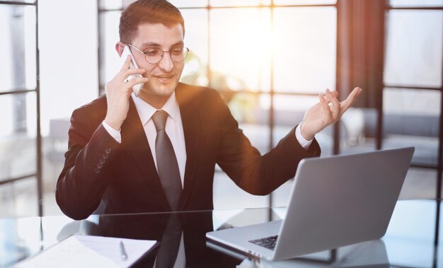 Portrait d'un jeune homme d'affaires sérieux dans un bureau décontracté appelant au téléphone tout en travaillant avec un ordinateur portable