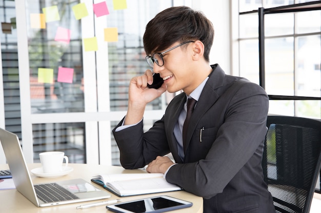 Portrait d'un jeune homme d'affaires souriant parlant sur son smartphone alors qu'il travaillait sur son ordinateur, assis dans son bureau au bureau portant un costume noir.