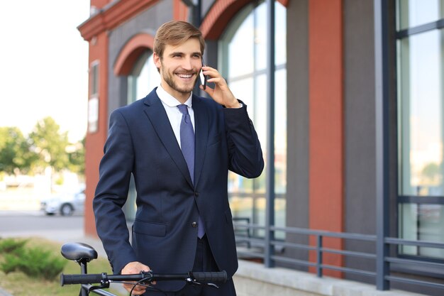 Portrait d'un jeune homme d'affaires souriant vêtu d'un costume parlant sur mobile debout à l'extérieur.