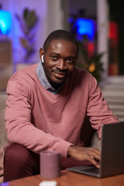 Photo portrait de jeune homme africain dans les écouteurs regardant la caméra alors qu'il était assis sur un canapé devant la table et en tapant sur un ordinateur portable