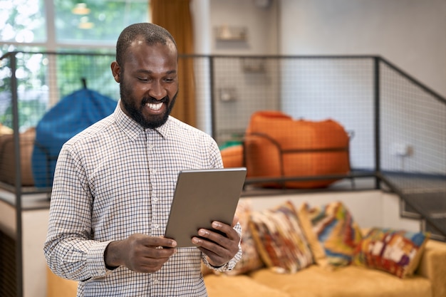 Portrait d'un jeune homme africain gai utilisant une tablette numérique en se tenant debout dans le bureau moderne