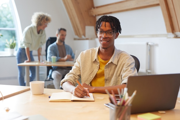 Portrait de jeune homme africain à lunettes assis sur son lieu de travail souriant à la caméra tout en prenant des notes dans son bloc-notes au bureau