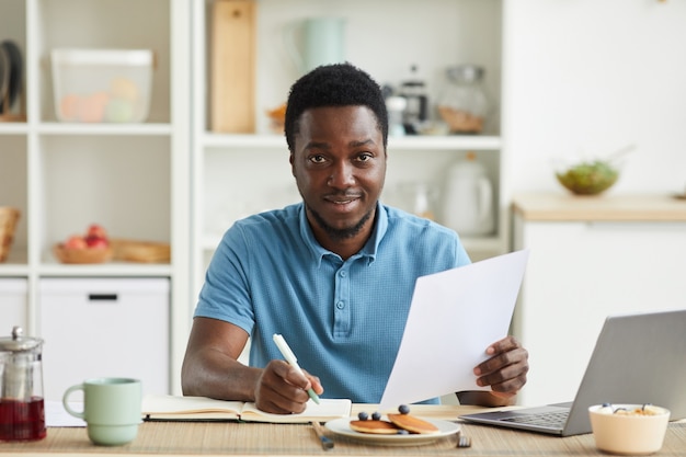 Portrait de jeune homme africain planifier sa journée et prendre des notes dans le bloc-notes alors qu'il était assis à la table dans la cuisine