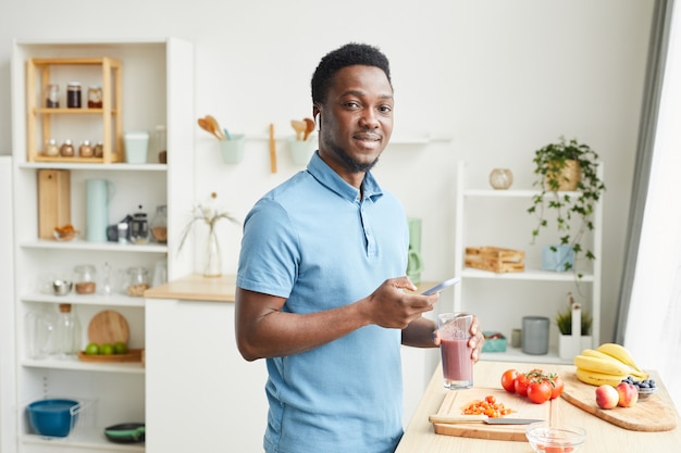 Portrait de jeune homme africain souriant tout en buvant du jus et en utilisant son téléphone portable dans la cuisine domestique