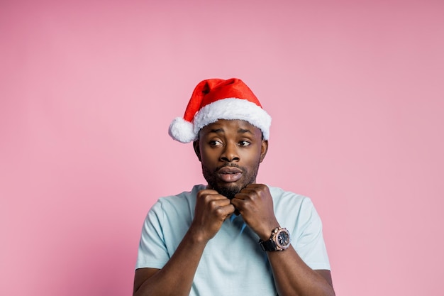 Portrait d'un jeune homme afro-américain effrayé et anxieux, gardant les mains sous le menton, portant un t-shirt blanc, un bonnet de Noel. L'étudiant se sent concerné avant l'examen. Expressions faciales humaines. Fond rose.