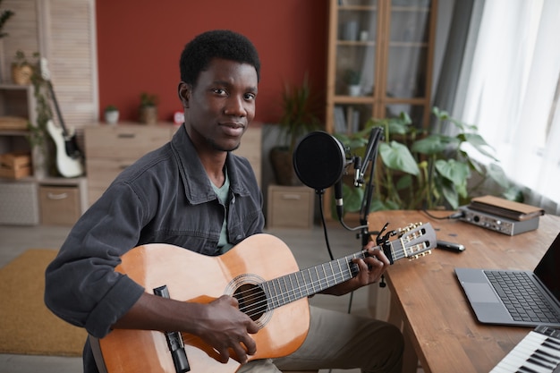 Portrait de jeune homme afro-américain jouant de la guitare et regardant la caméra alors qu'il était assis par microphone en studio d'enregistrement à domicile, espace copie