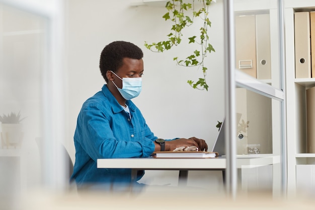 Portrait de jeune homme afro-américain portant un masque tout en travaillant dans une cabine au bureau post pandémique