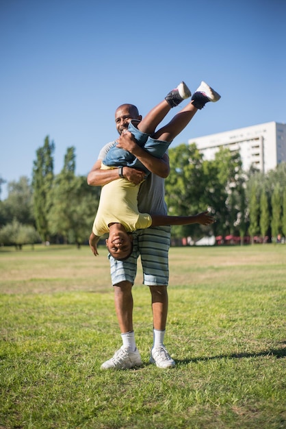 Portrait de jeune homme afro-américain tenant un garçon. Papa souriant en short debout sur l'herbe avec un fils heureux à l'envers. Moments de bonheur et concept de repos actif