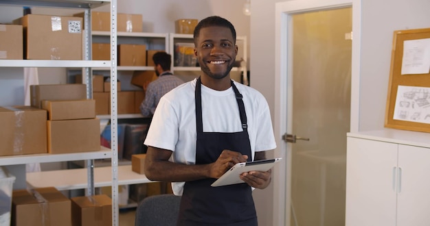 Portrait d'un jeune homme afro-américain utilisant une tablette numérique dans un entrepôt faisant l'inventaire souriant à la caméra