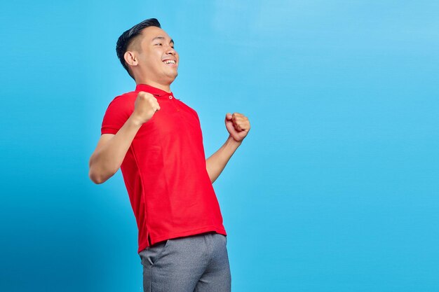 Portrait d'un jeune homme asiatique excité en chemise rouge célébrant le succès avec des expressions faciales joyeuses sur fond bleu