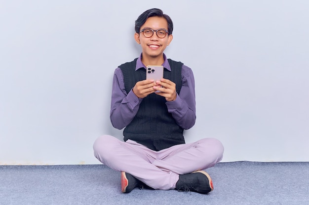 Portrait d'un jeune homme asiatique heureux utilisant un téléphone portable assis sur le sol avec les jambes croisées isolé sur fond blanc