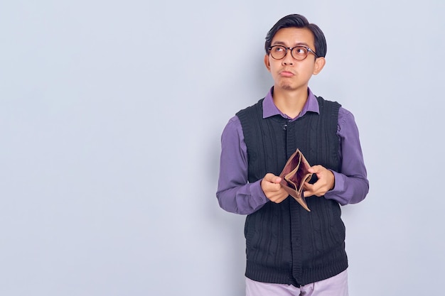 Portrait d'un jeune homme asiatique malheureux portant une chemise et un gilet montrant un portefeuille vide isolé sur fond blanc