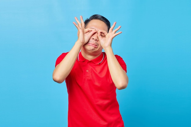 Portrait d'un jeune homme asiatique montrant un signe ok avec les deux doigts et couvrant les yeux sur fond bleu