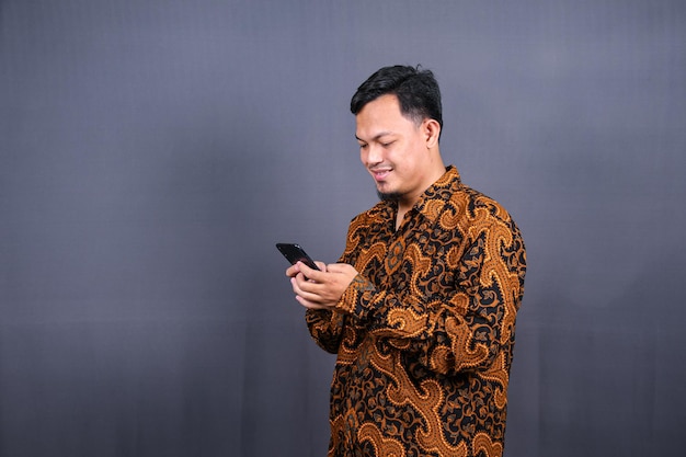 Portrait d'un jeune homme asiatique portant du batik à l'aide d'un téléphone portable sur fond gris