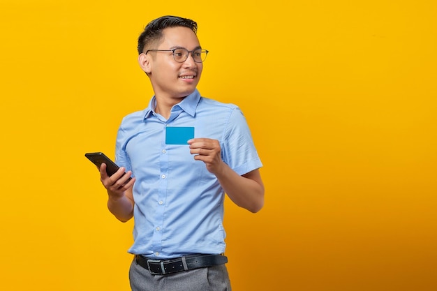 Portrait d'un jeune homme asiatique souriant asiatique dans des verres tenant un téléphone portable et une carte de crédit isolé sur fond jaune concept d'homme d'affaires et d'entrepreneur