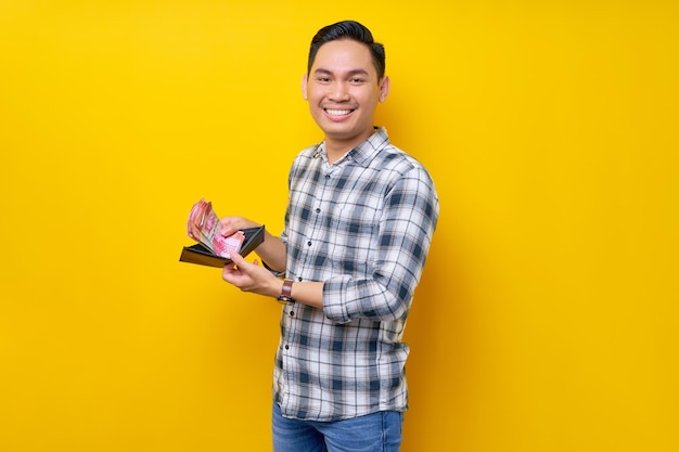 Portrait d'un jeune homme asiatique souriant portant une chemise à carreaux tenant un portefeuille plein d'argent liquide en billets de roupie à la main isolé sur fond jaune concept de mode de vie des gens