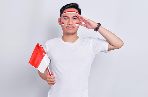 Portrait d'un jeune homme asiatique souriant tenant un drapeau indonésien et montrant un geste de respect tout en célébrant le jour de l'indépendance indonésienne le 17 août isolé sur fond blanc