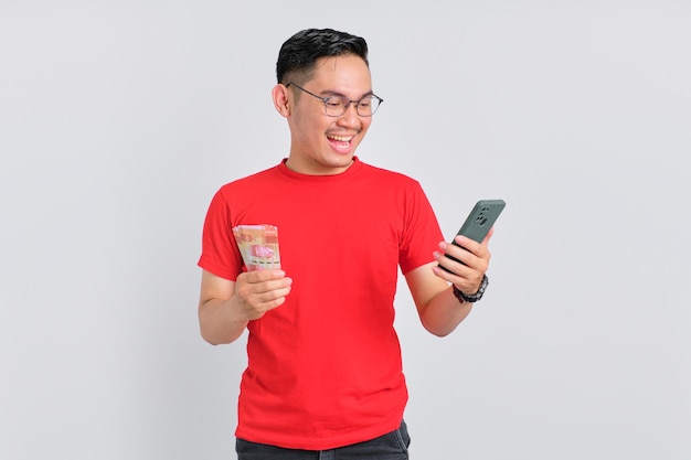 Portrait d'un jeune homme asiatique souriant utilisant un téléphone portable et tenant des billets de banque isolés sur fond blanc