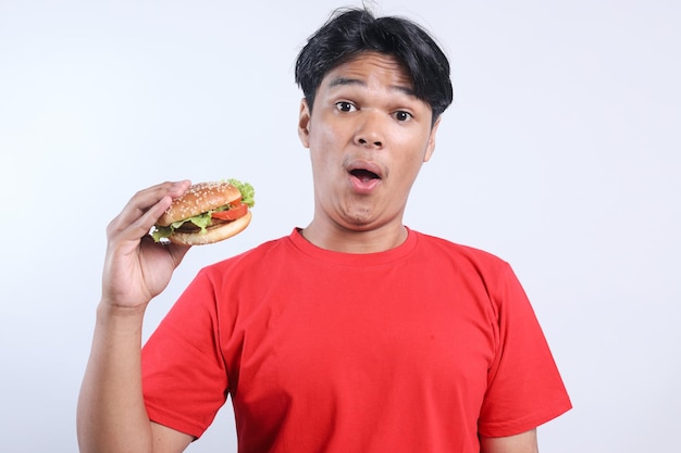 Portrait d'un jeune homme asiatique surpris levant la main tout en tenant un hamburger isolé sur un fond blanc