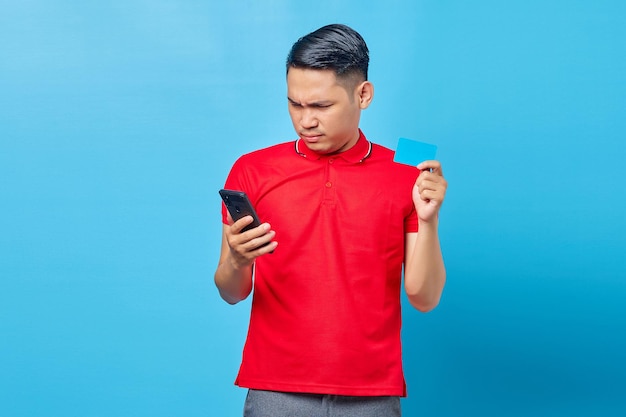 Portrait d'un jeune homme asiatique surpris tenant un téléphone portable et montrant une carte de crédit isolée sur fond bleu