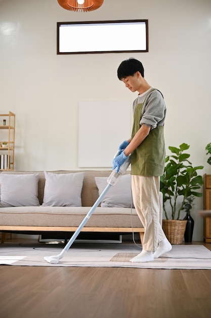 Portrait d'un jeune homme asiatique utilisant un aspirateur-balai pour nettoyer le tapis dans le salon