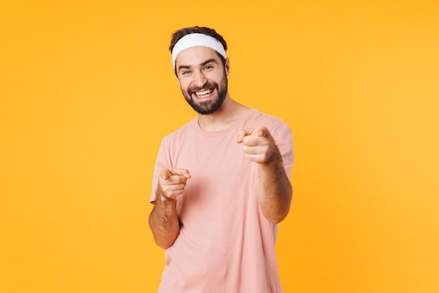 Portrait d'un jeune homme athlétique musclé en t-shirt souriant et pointant du doigt isolé sur un mur jaune