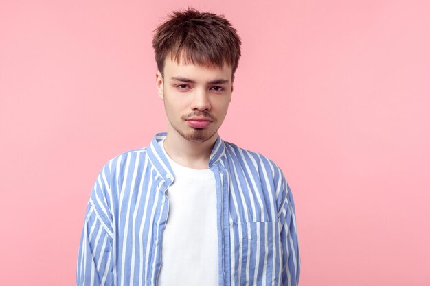 Portrait d'un jeune homme aux cheveux bruns malheureux avec une petite barbe et une moustache en chemise rayée décontractée debout regardant tristement l'humeur déprimée de la caméra prise de vue en studio à l'intérieur isolée sur fond rose