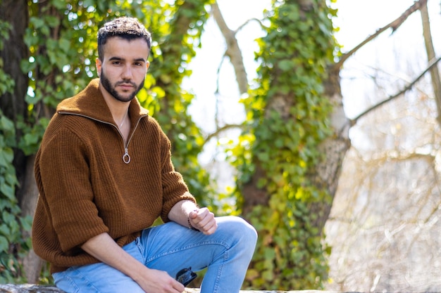 Portrait d'un jeune homme barbu attrayant assis dans un environnement naturel Beau jeune homme assis à l'extérieur en regardant la caméra