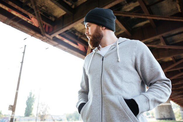 Photo portrait d'un jeune homme barbu décontracté au chapeau debout sous un pont urbain et à l'écart