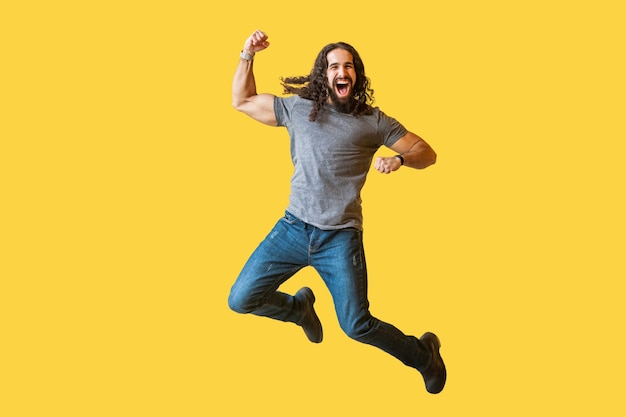 Portrait d'un jeune homme barbu joyeux et joyeux aux longs cheveux bouclés en t-shirt gris décontracté sautant et célébrant sa victoire avec un visage excité étonné. studio intérieur tourné isolé sur fond jaune.