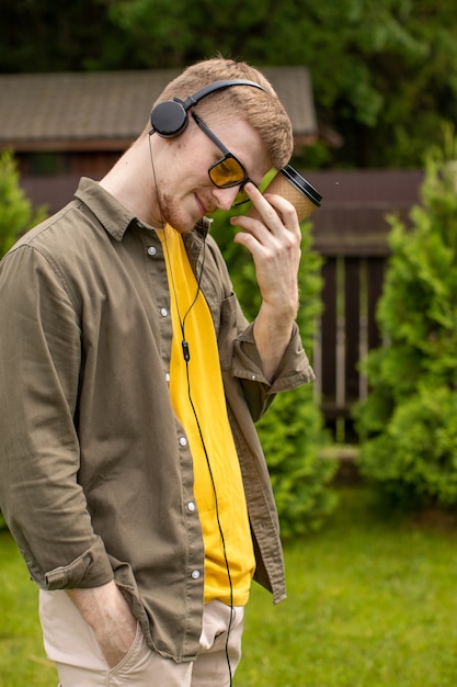 Portrait de jeune homme barbu à lunettes jaunes et t-shirt jaune habillé avec désinvolture debout avec une tasse en papier jetable en écoutant attentivement de la musique en ligne avec des écouteurs sur fond vert