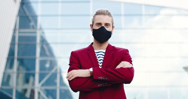 Portrait de jeune homme beau caucasien en masque regardant la caméra avec le visage souriant et croisant les mains à l'extérieur. Élégant mâle beau à la rue. Concept de pandémie de coronavirus. Distance sociale.