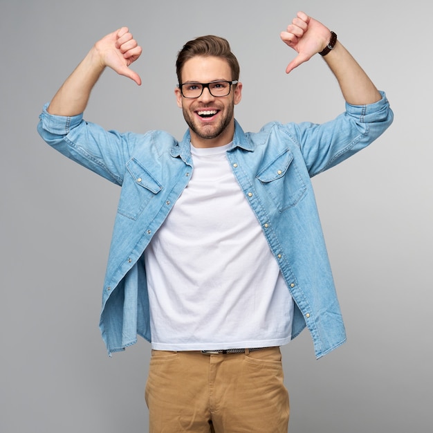 Portrait de jeune homme beau en chemise de jeans montrant à lui-même par de gros pouces debout sur un mur léger