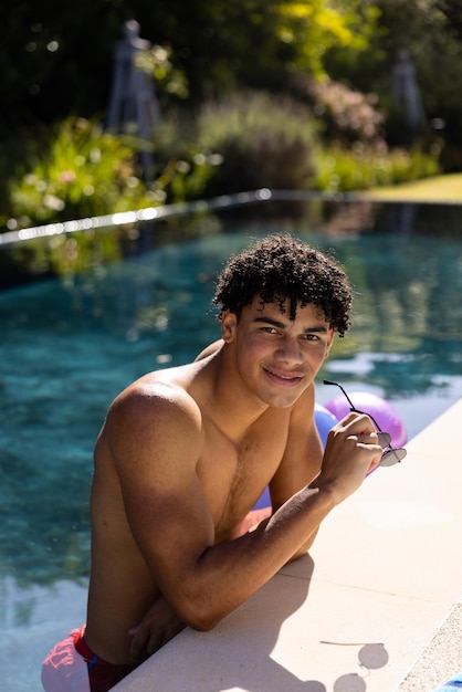 Portrait d'un jeune homme biracial torse nu tenant des lunettes de soleil souriant et penché au bord de la piscine. Espace de copie, inchangé, beau, dreadlocks, style de vie, plaisir, concept d'activités d'été et de week-end.