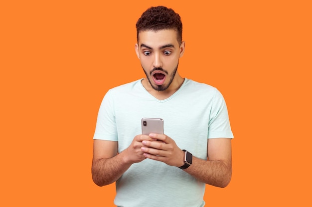 Portrait de jeune homme brune avec barbe en t-shirt blanc exprimant son étonnement tout en utilisant un téléphone portable debout avec la bouche ouverte et de grands yeux choqués tourné en studio intérieur isolé sur fond orange