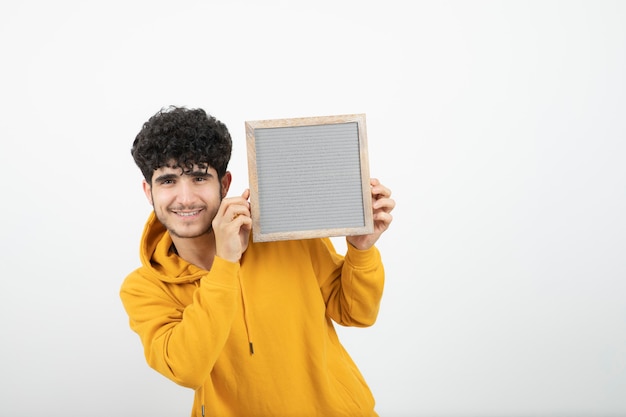 Portrait d'un jeune homme brune debout et tenant un cadre gris.