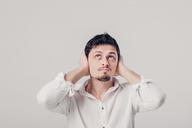 Portrait d'un jeune homme Brunet en chemise blanche couvre ses oreilles avec ses mains sur un fond gris. Je ne veux rien entendre. Lumière douce