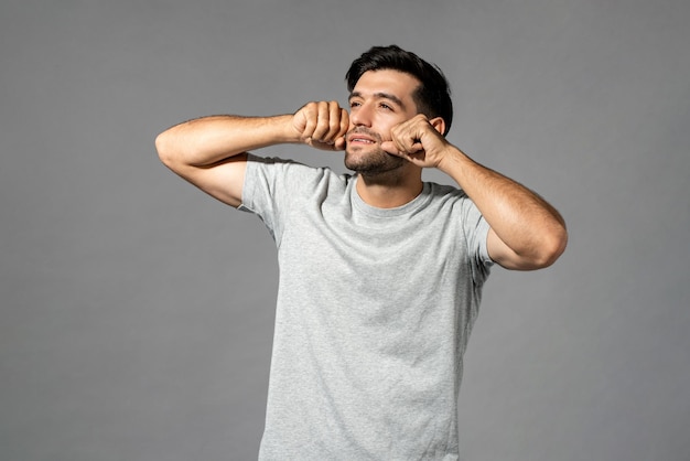 Portrait d'un jeune homme caucasien beau et endormi, se frottant les yeux après le réveil et regardant loin dans un fond de studio gris clair isolé