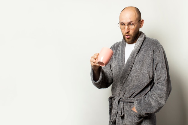 Portrait d'un jeune homme chauve avec une barbe dans une robe de chambre et des verres à la surprise dans une tasse de café