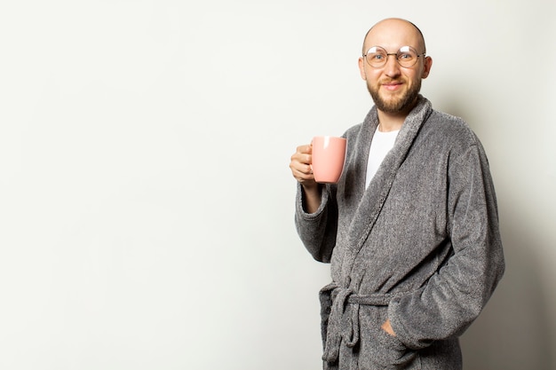 Portrait d'un jeune homme chauve avec une barbe dans une robe de chambre et des verres tenant une tasse de café chaud